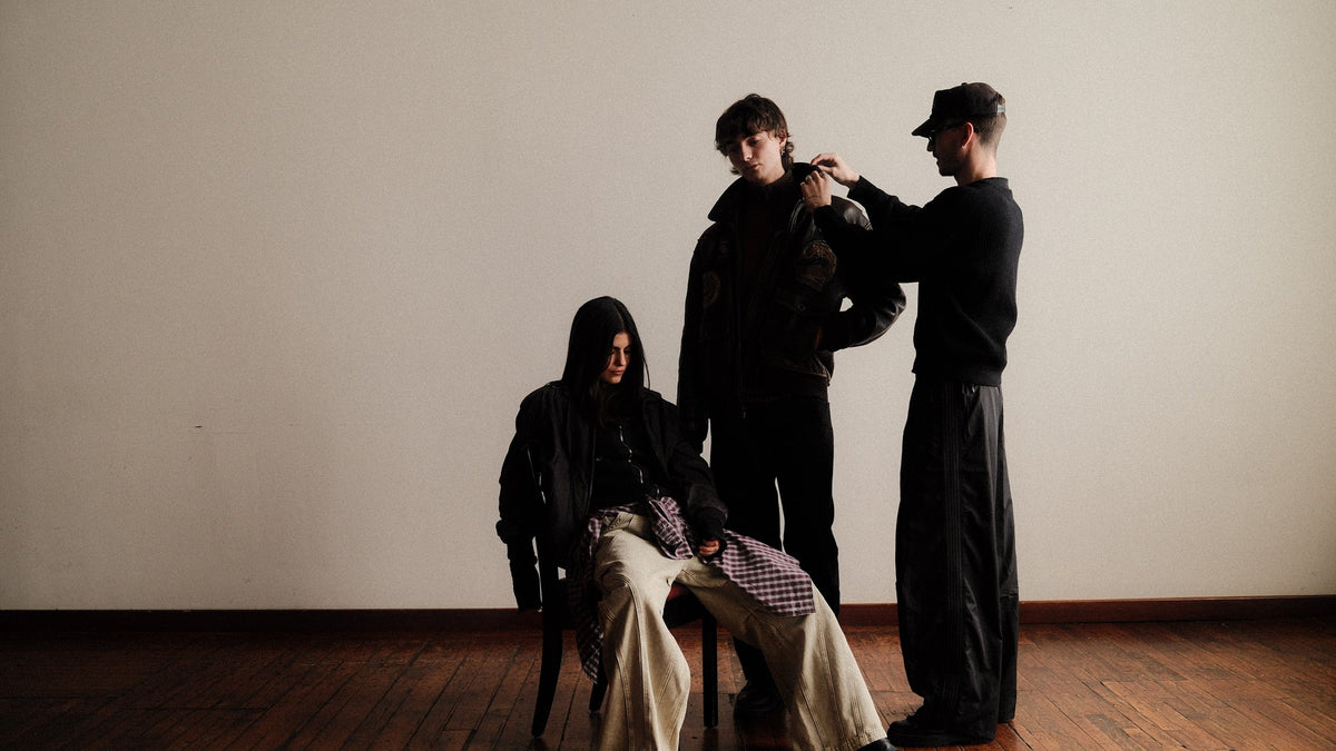 Three people in dark clothing pose in a minimalist studio with a plain off-white wall and wooden floor. One person sits on a chair while the other two stand, with the standing figure on the right adjusting the clothing of the central figure.